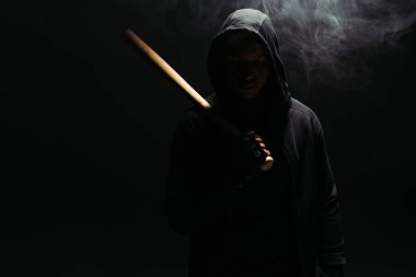 Silhouette of african american hooligan in hood holding baseball bat on black background with smoke  clipart