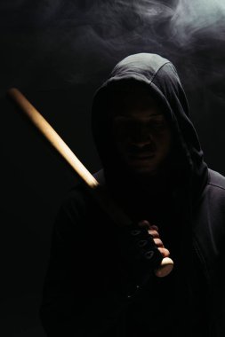 Silhouette of african american bandit in hood and glove holding baseball bat on black background with lighting and smoke  clipart