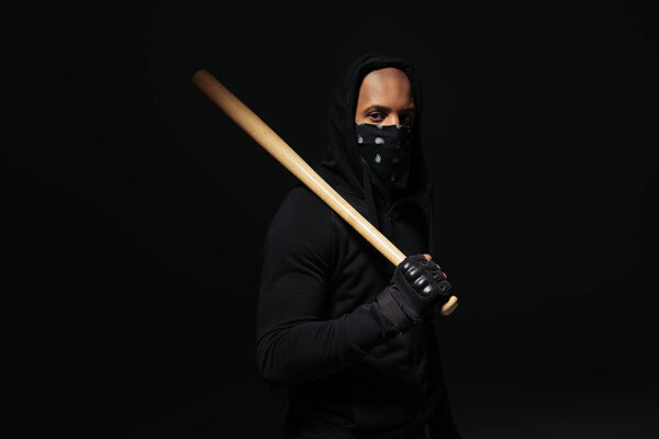 African american hooligan with mask on face holding wooden baseball bat isolated on black 