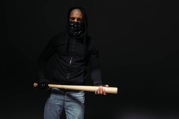 African american hooligan in gloves and mask on face holding baseball bat isolated on black