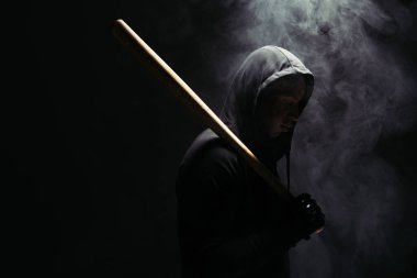 Side view of african american hooligan holding baseball bat in lighting on black background with smoke  clipart