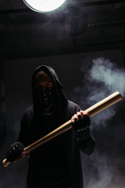 Silhouette of african american vandal with mask on face holding baseball bat on black background with smoke  clipart
