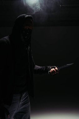 Silhouette of african american robber in mask and hoodie holding knife on black background with smoke 
