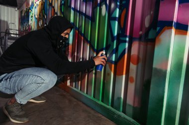 Side view of african american vandal in hood drawing graffiti on wall 