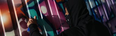 Side view of african american vandal in mask drawing graffiti on wall at night, banner 
