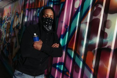 African american vandal with mask on face holding spray paint near graffiti on wall  clipart
