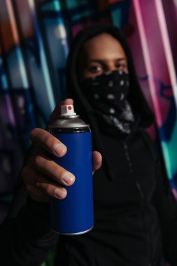 Blurred african american vandal with mask on face holding spray paint near graffiti on urban street 