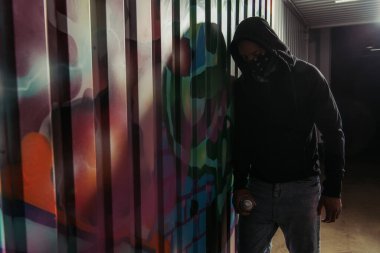 African american vandal holding spray paint near graffiti on wall 