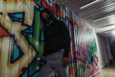 African american vandal in hood holding spray paint near graffiti on wall 