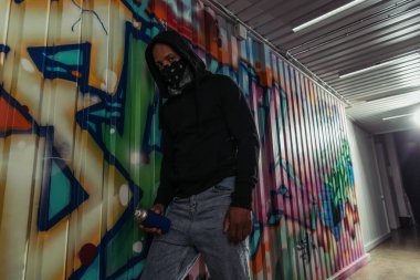 African american vandal in hood holding spray paint near graffiti on wall  clipart