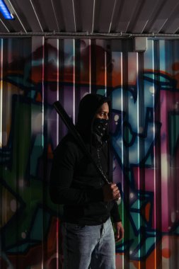Side view of african american hooligan in mask and hoodie holding baseball bat near graffiti on wall  clipart