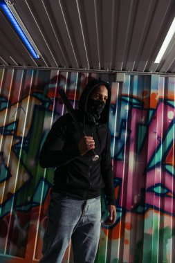African american vandal in mask holding baseball bat near graffiti on wall 