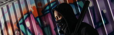 Side view of african american bandit in mask holding baseball bat near graffiti on wall, banner 