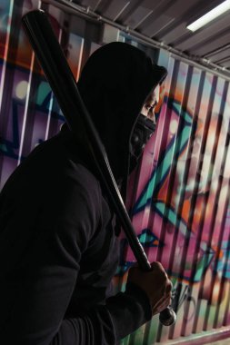 Duvarda duvar yazısı olan beyzbol sopası tutan maskeli Afro-Amerikan vandalının yan görüntüsü 