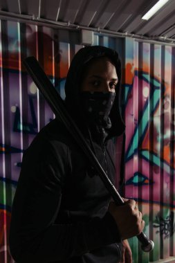 African american hooligan with baseball bat looking at camera near graffiti on wall 