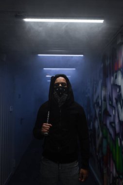 African american hooligan in mask holding baseball bat and looking at camera near lighting and graffiti 
