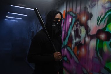 African american hooligan with covered face holding baseball bat near graffiti in garage 
