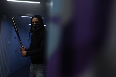 Yüzünde maske olan Afrikalı Amerikalı holigan garajdaki bulanık duvarın yanında beyzbol sopası tutuyor.