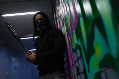 Low angle view of african american hooligan with covered face holding baseball bat near graffiti 