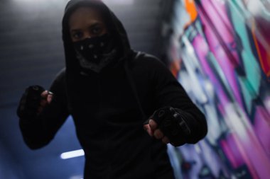 Low angle view of blurred african american hooligan in gloves and mask standing in fighting pose in garage 