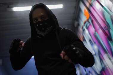 Low angle view of african american bandit in mask standing in fighting pose near graffiti in garage 