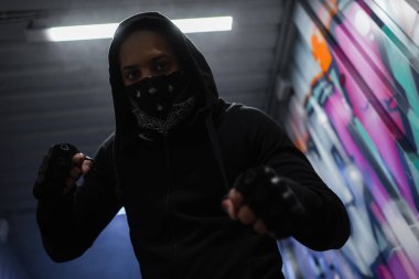 Low angle view of african american bandit in mask standing in fighting pose near graffiti in garage  clipart