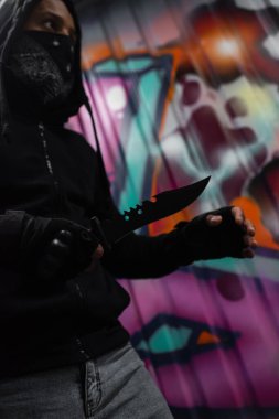 Low angle view of african american bandit in mask holding knife near graffiti outdoors 
