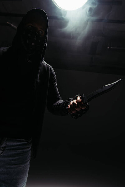 Silhouette of african american hooligan with obscure face holding knife on black background with smoke 