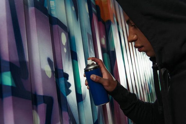 Side view of african american hooligan in hoodie drawing graffiti on wall on urban street 