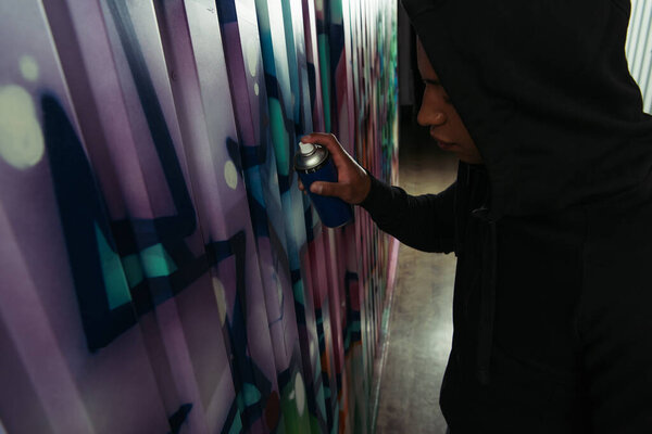 Side view of african american vandal with spray paint drawing graffiti on wall 