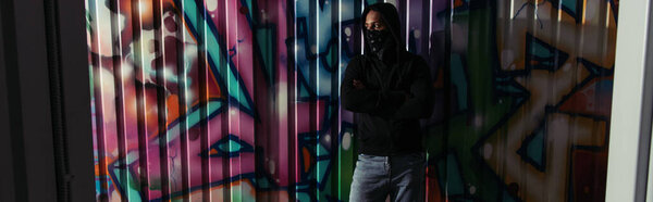African american hooligan in hoodie and face mask standing near graffiti on wall, banner 