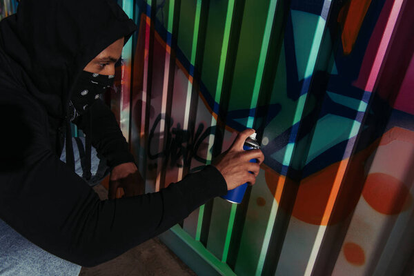 Side view of african amercan hooligan in mask drawing graffiti on wall at night 