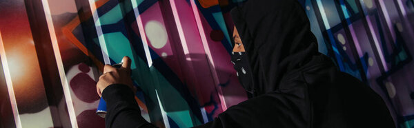 Side view of african american vandal in mask drawing graffiti on wall at night, banner 