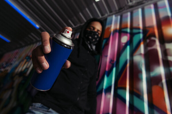 Low angle view of blurred african american vandal holding spray paint near graffiti on wall 
