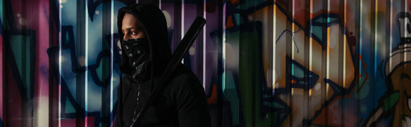 Африканский американский хулиган в маске держит бейсбольную биту возле граффити на стене на городской улице, баннер 