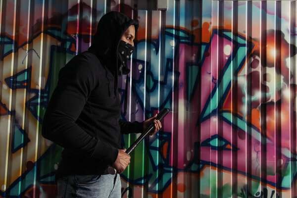 Side view of african american vandal holding baseball bat near graffiti on wall on street 