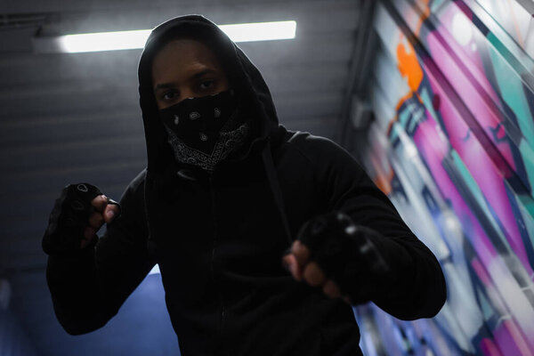 Low angle view of african american bandit in mask standing in fighting pose near graffiti in garage 