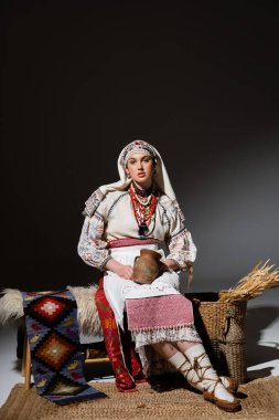 full length of young ukrainian woman in traditional clothing with ornament holding clay pot while sitting on bench on black