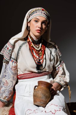 pretty ukrainian woman in traditional clothing with ornament holding clay pot on black