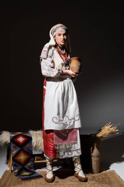 full length of pretty  ukrainian woman in traditional clothing with ornament holding clay pot while standing on black
