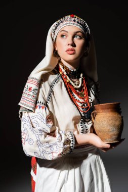 portrait of pretty ukrainian woman in traditional clothing with ornament holding clay pot on black clipart