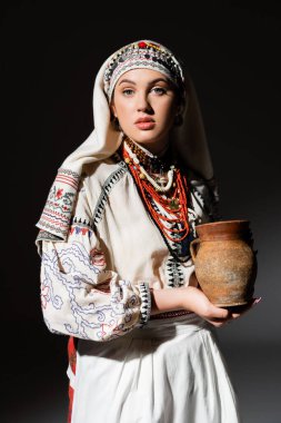 portrait of young ukrainian woman in traditional clothing with ornament holding clay pot on black