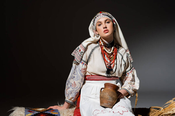 young ukrainian woman in traditional clothing with ornament holding clay pot and sitting on bench on black