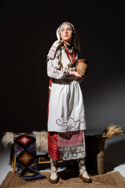 full length of young ukrainian woman in traditional clothing with ornament holding clay pot while standing on black