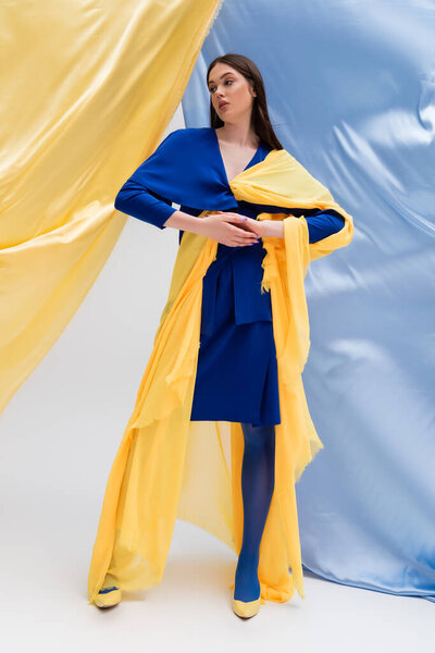full length of young ukrainian model in color block dress posing near blue and yellow curtains 