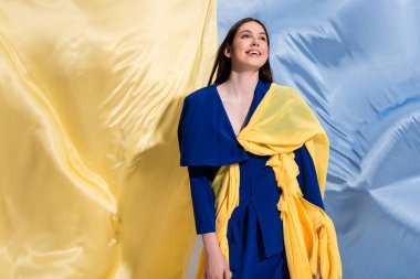 cheerful ukrainian woman in color block clothing posing near blue and yellow flag