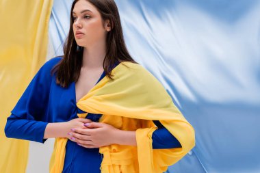 patriotic ukrainian young woman in stylish clothing posing near blue and yellow flag 