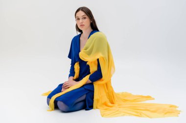 full length of patriotic ukrainian young woman in blue and yellow outfit sitting on grey