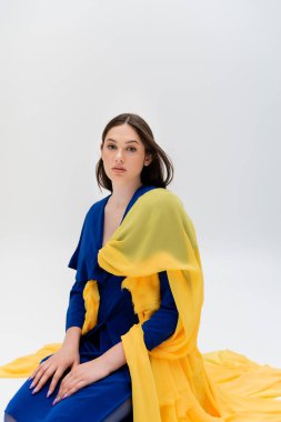 patriotic ukrainian young woman in blue and yellow outfit sitting and looking at camera on grey 