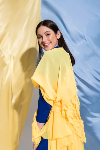happy ukrainian woman in color block clothing posing near blue and yellow fabric